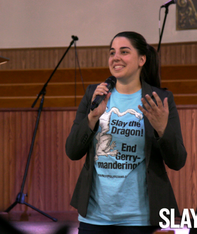 Katie Fahey wearing a t-shirt that says "Slay the Dragon, End Gerrymandering" while speaking in front of a group of people
