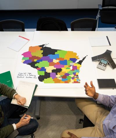 A bird's eye view of a map of Wisconsin on a table with district lines drawn and two people sitting next to it discussing.