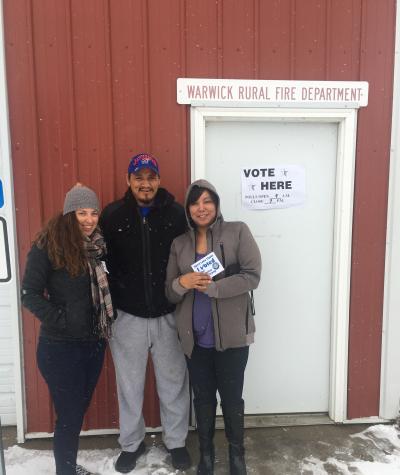 Three people outside a building in the snow. One of the women holds a sign that says "I Voted". A sign on the door to the building says "Vote Here".