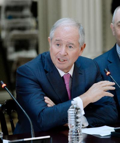 Stephen Schwarzman speaks into a microphone while sitting at a table while President Trump looks on