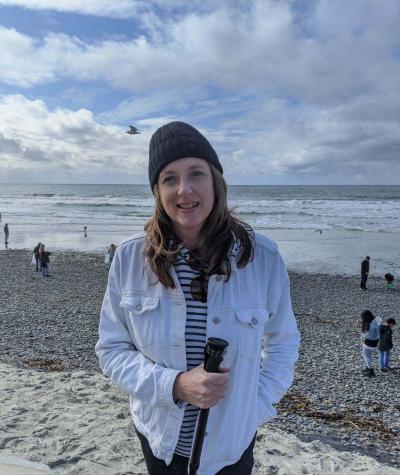 Maria Romo wearing a winter hat standing on a beach with the water behind her