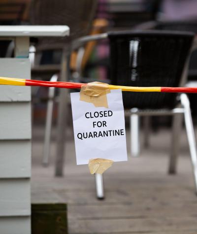 Close up of a sign hanging in front of an empty outdoor seating area of a cafe that says "Closed for Quarantine"