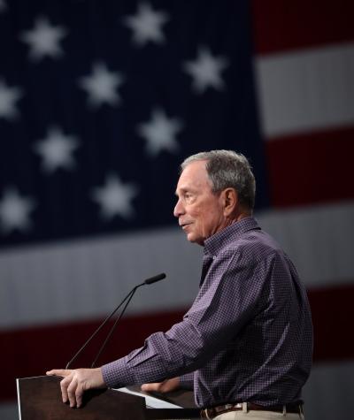 Michael Bloomberg speaking at a podium with an American flag in the background