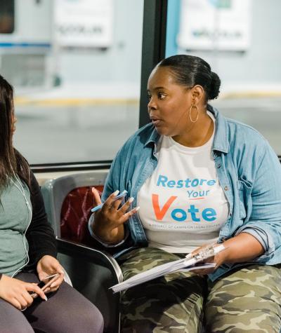 A person wearing a Restore Your Vote t-shirt talking to another person on a bench