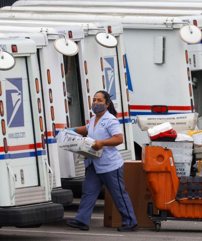A woman wearing a mask carries a crate to the back of a postal truck