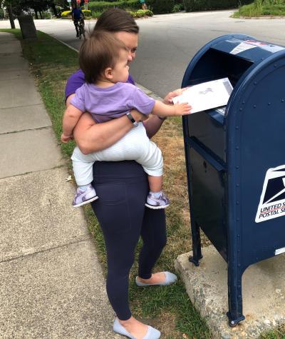 Ruth Greenwood placing her ballot in a blue USPS mailbox while holding her daughter.