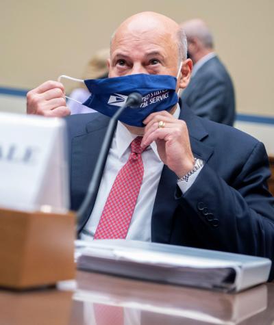Louis DeJoy seated behind a desk removing a dark blue face mask with the Postal Service logo on it
