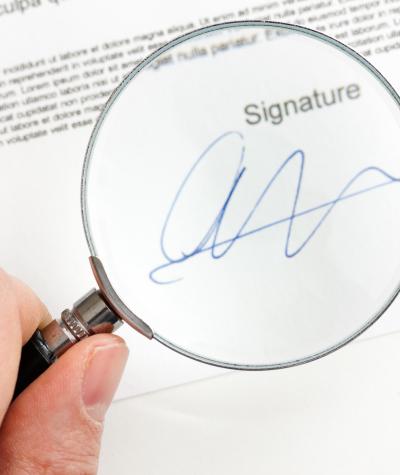 Close up of a person's hand holding a magnifying glass over a signature on a document