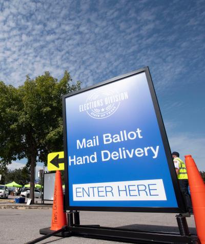 An election official in a neon yellow vest stands next to a large blue sign that says "Mail Ballot Delivery, Enter Here"