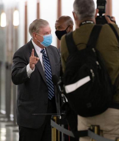 Lindsey Graham wearing a mask and gesturing while speaking to a densely packed group of reporters