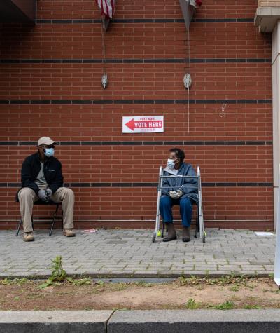 A man and elderly woman wearing masks sit on chairs on a sidewalk with a "Vote Here" sign on the building behind them.