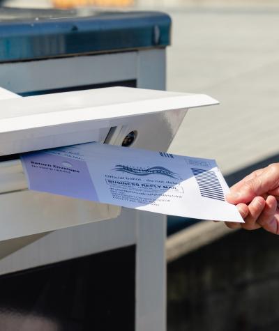 A hand holding a ballot inserts it into a slot in a drop box.