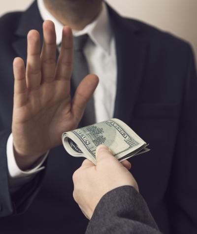 A man in a suit holding up his hand to refuse a wad of money being handed to him by another man off-camera