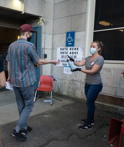 A woman dispenses hand sanitizer to a man and woman outside the open door of a building with "Vote Here" signs on the outside.