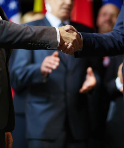 Two men in suits shaking hand with people clapping in the background
