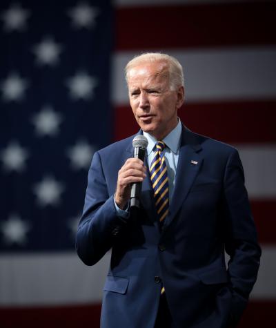 Joe Biden speaking while holding a microphone in front of a large American flag