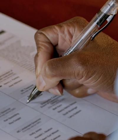 A woman's hand filling out a paper ballot with a pen.
