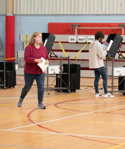 A woman wearing a mask and carrying a ballot walks across the floor of a gym while being directed by another woman who is an election official.