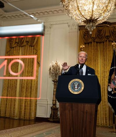 Joe Biden giving a speech at a podium with a large red "ON AIR" sign hanging in front of him