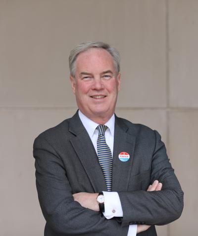Trevor Potter posing with his arms folded in front of a wall and wearing an I Voted sticker on his lapel