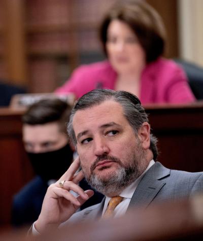 Ted Cruz resting his head on his fingers while sitting at a desk with several people out of focus in the background behind him.