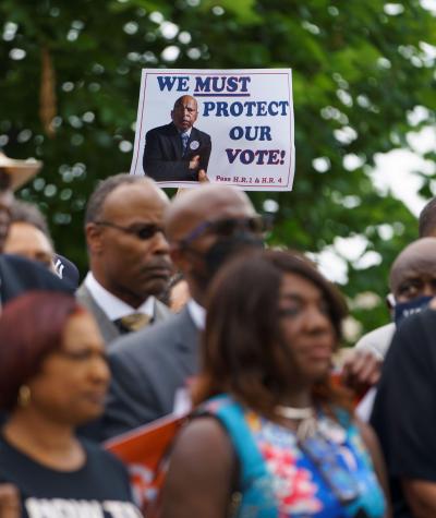 A sign with John Lewis's picture on it saying "We must protect our right to vote" is held above the heads of a crowd standing in front of some trees