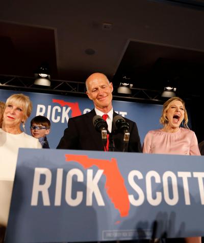Rick Scott standing behind a podium with his name and an outline of the state of Florida on the front surrounded by several other people