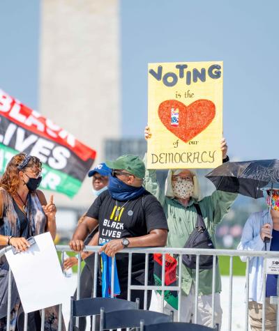 People with sign stand behind a barricade in front of the Washington Monument. One sign reads "Voting is the heart of our democracy". A man in the background carries a Black Lives Matter Flag.
