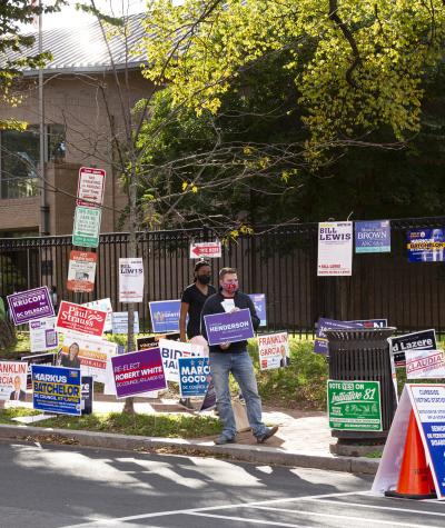 A person holds a sign while sanding among other candidate signs on a sidewalk corner