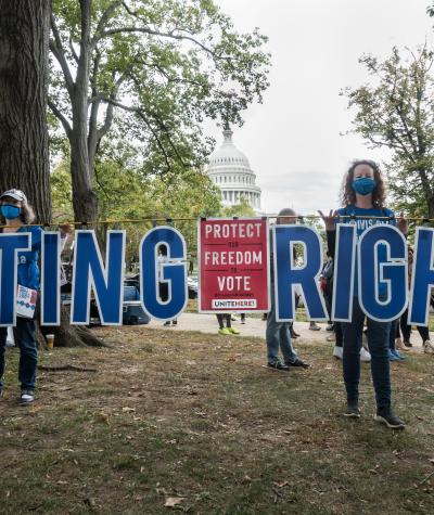 Two women stand among trees in front of the Capitol Building holding large letters that say "Voting Rights". A sign in between them reads "Protect our freedom to vote"