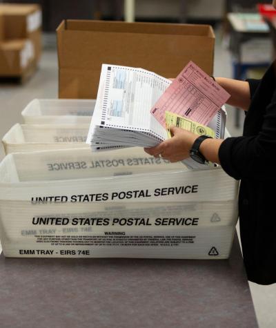 A woman lifts a stack of ballots out of a postal crate.