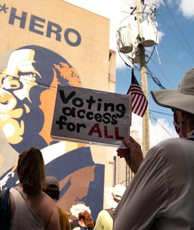 A woman with her back to the camera holds a sign which says "Voting access for ALL", while standing with others in front of a large mural of John Lewis.