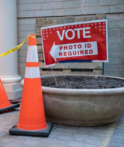 A sign which says "Vote, Photo ID Required" next to some caution tape and construction cones.
