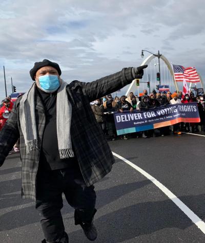 A man wearing a surgical mask walks out in front of a group of people crossing a bridge with his finger pointing forward. The people are holding an American flag and carrying a sign that says "Deliver for Voting Rights."