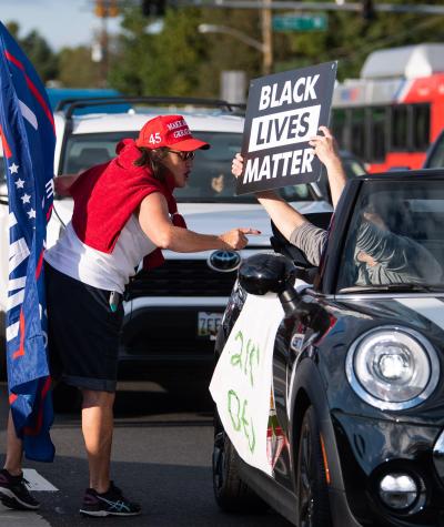 A woman wearing a "Make America Great Again" hat points her finger and yells at a person driving by in a car holding up a "Black Lives Matter" sign.