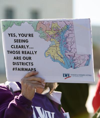 A woman's hand holding a sign with a map of Maryland which says "Yes, you're seeing clearly...those really are our districts. #fairmaps"