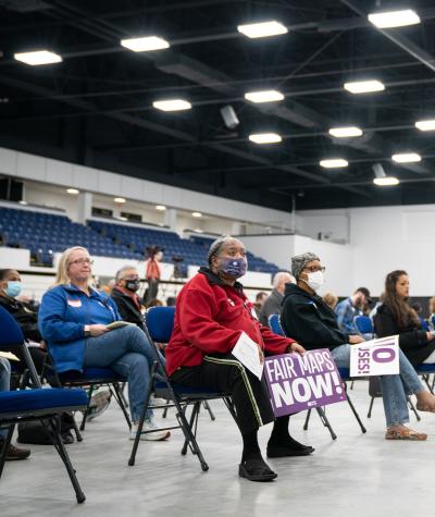A woman seated in the front row of a group of folding chairs holds a sign which reads "Fair maps now". Others holding signs are seating next to her.