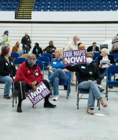 People sit on folding chairs in a gymnasium holding sign that say "Fair Maps NOW!"