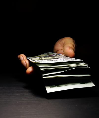 A hand emerges out of the dark holding a stack of $100 bills.