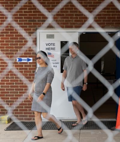 An open door of a brick building with two people exiting and a "Vote Here' sign on it all overlaid with an out-of-focus chain link fence.