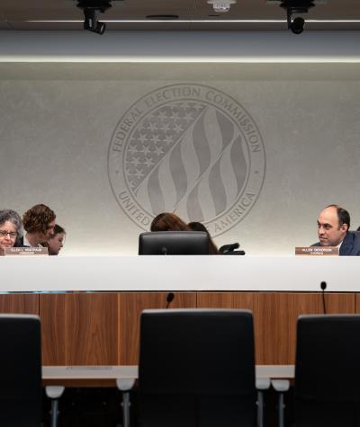 People sit behind a large desk with microphone in front of them and a large logo behind at the Federal Election Commission.