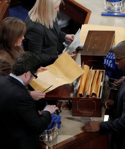 A top-down view of people in Congress opening a box containing manilla envelopes
