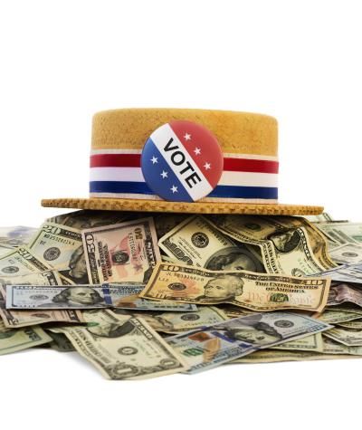 A straw hat with a red white and blue ribbon and a "Vote" button on it sitting on top of a pile of money.
