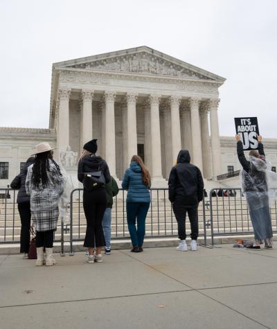 People stand a long a fence in front of the Supreme Court. One woman wearing a poncho holds up a sign which says "It's about US".