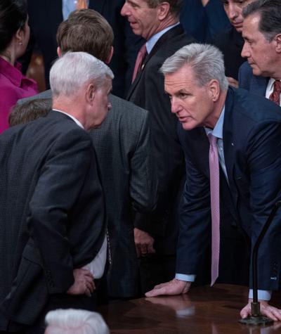Kevin McCarthy leaning forward with his hands on a desk speaking with another man with other members of Congress around them