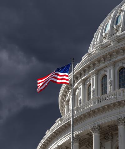 A close up of the dome of the U.S. Capitol Building with an American flag flying and a dark cloudy sky in the background