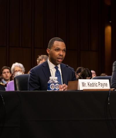 Kedric Payne speaking into a microphone and seated at a table next to another man with audience members behind them.