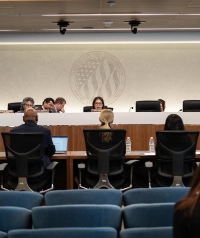 FEC commissioners sit behind a desk facing five people seated in chairs at a desk in front of them