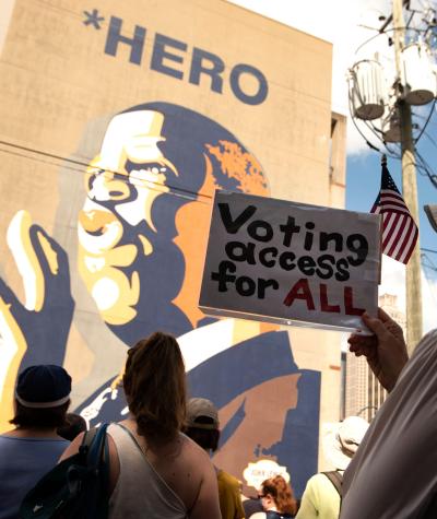 A woman holds a sign which says "Voting Access for All" while standing in front of a large mural of John Lewis.