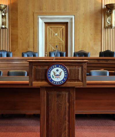 A podium with a microphone with the Senate seal on the front in front of two rows of chairs behind desks.